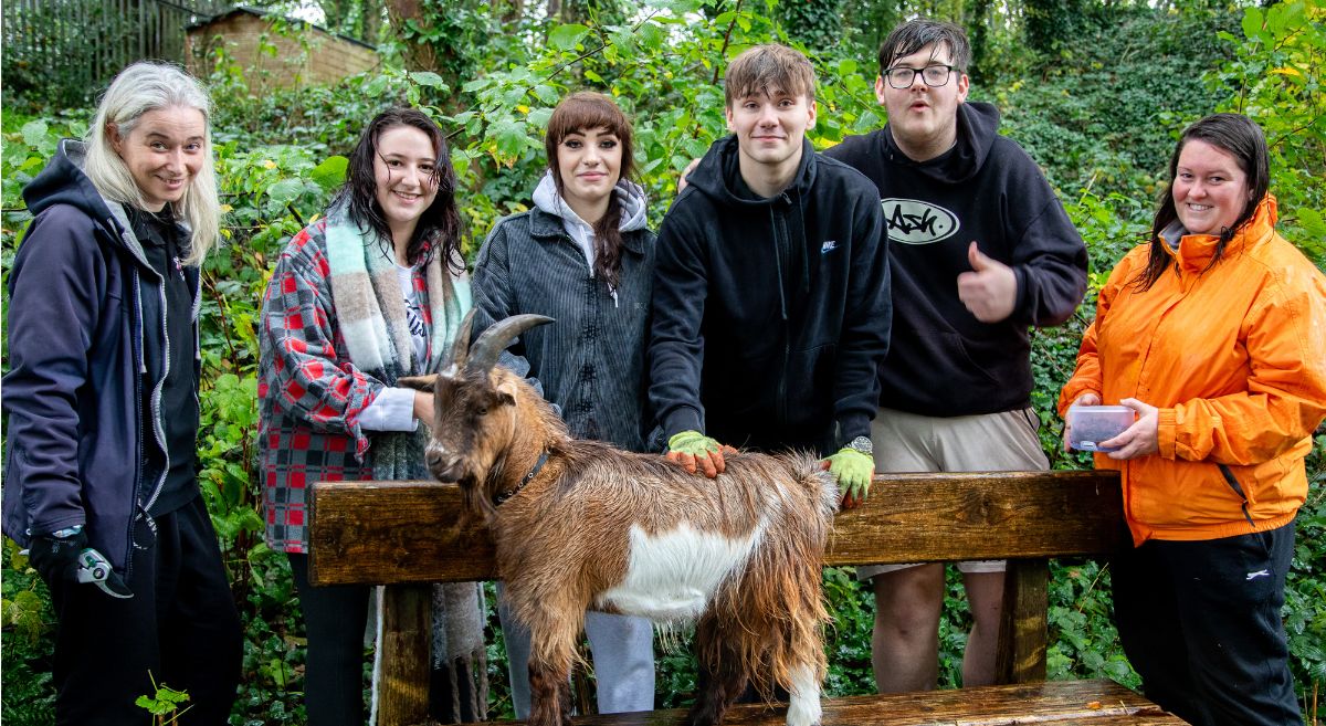 Six people dressed in outside clothing standing behind a smally pigmy goat which is standing on a bench